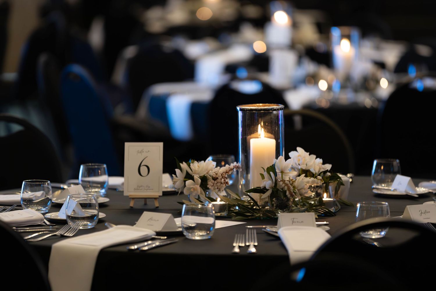Elegant wedding reception table with black linen, candle centerpiece, white flowers, and table number six in a dimly lit ballroom.