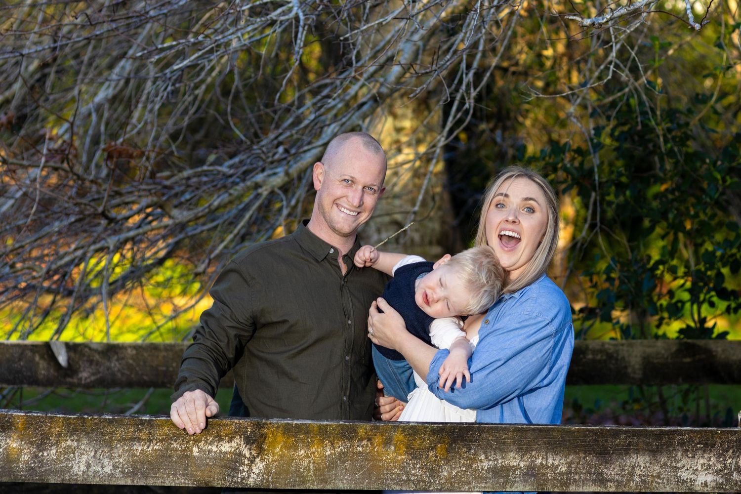 A happy young family with a baby poses outdoors near a wooden fence with autumn trees in the background.