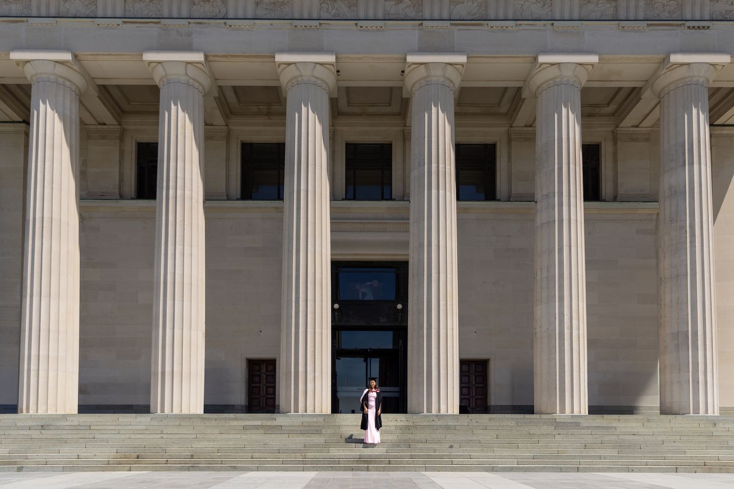 Person standing on steps of a neoclassical building with tall Ionic columns and grand stone facade.