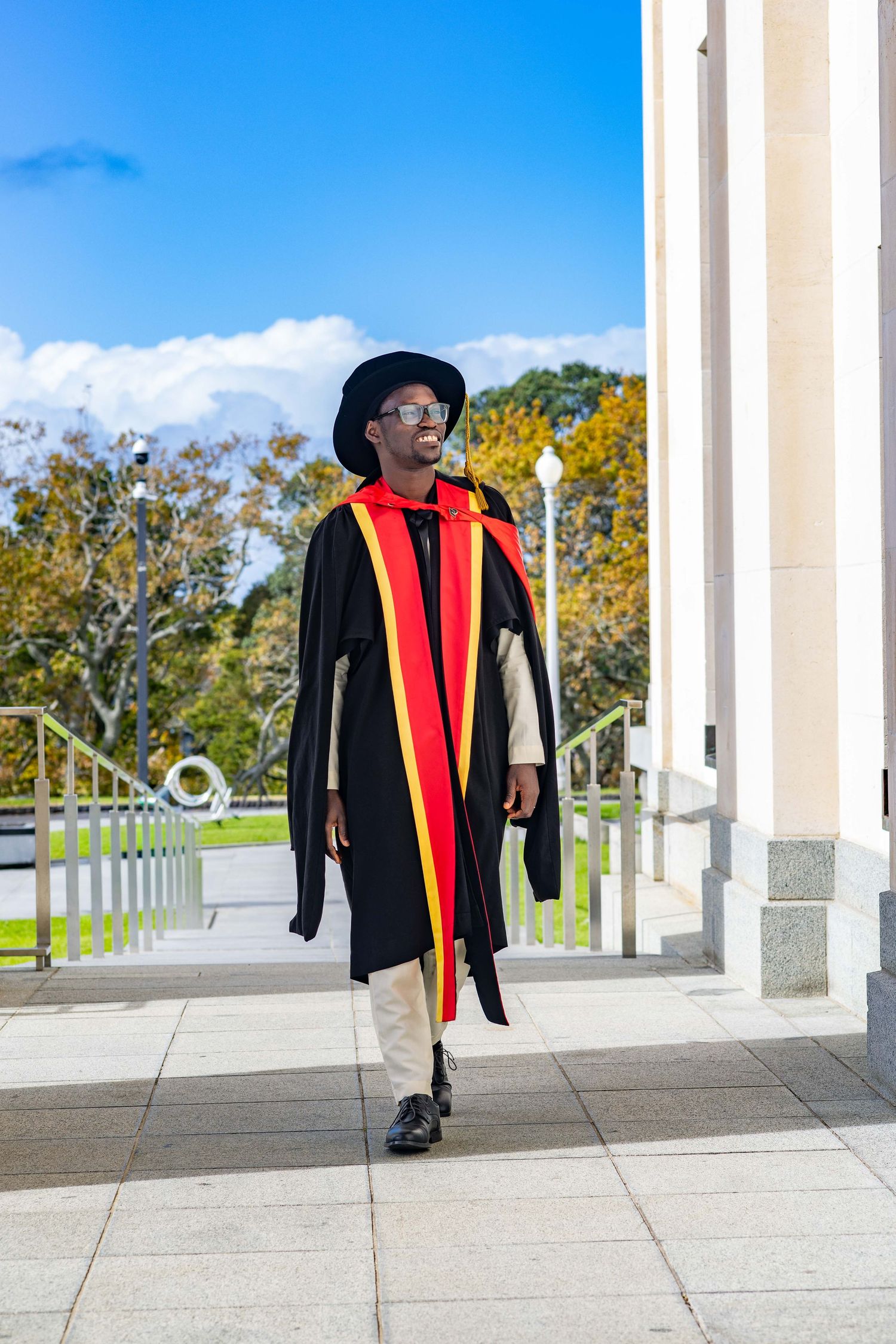 A young Black man in graduation gown, red and yellow sash, and mortarboard hat walks confidently outdoors.