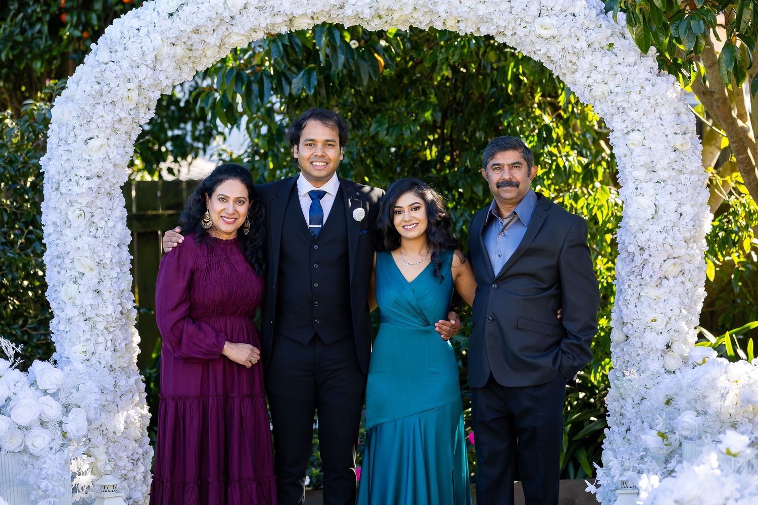 Family of four posing under a white floral arch at a wedding, dressed in formal attire outdoors.