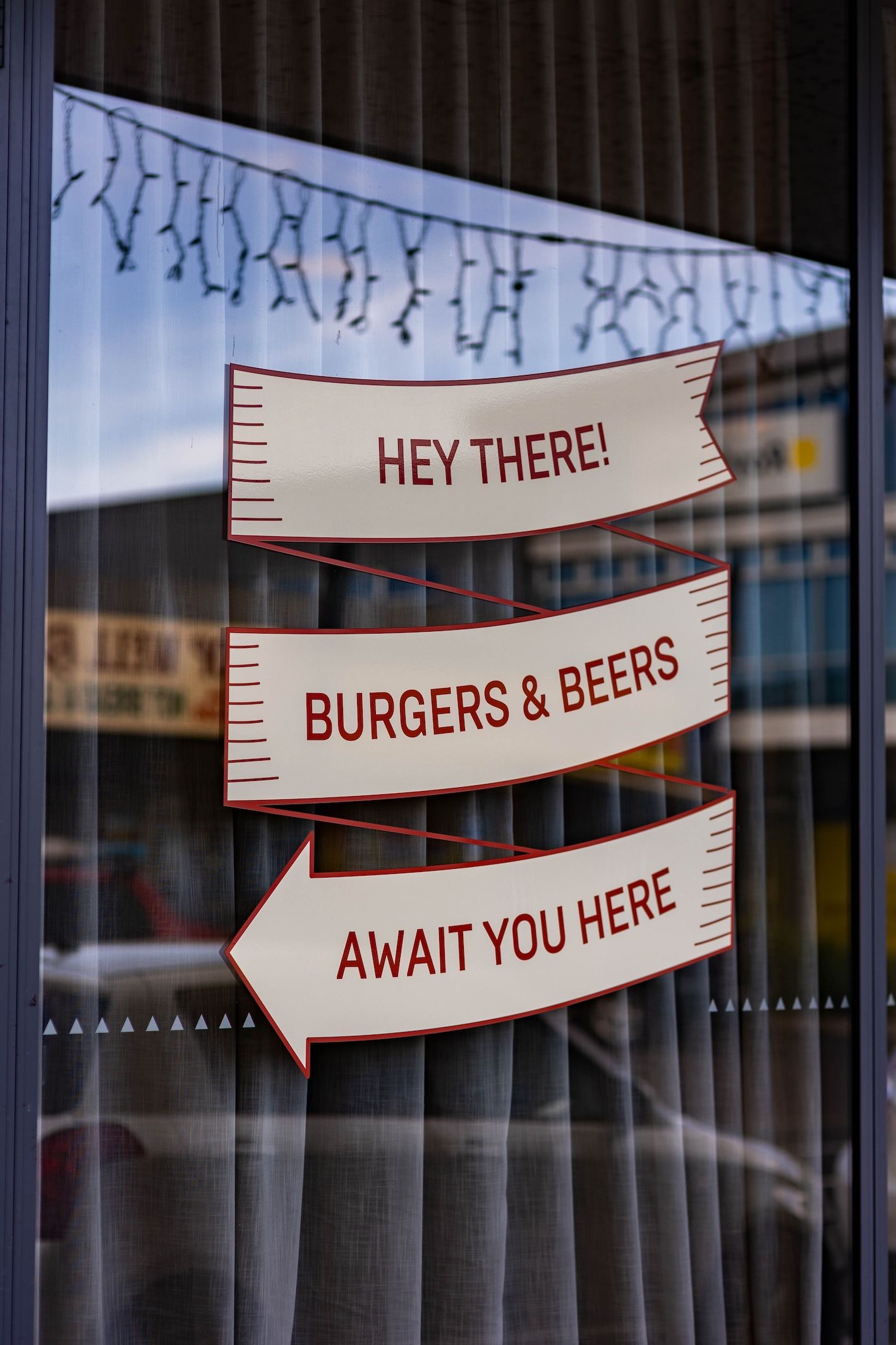 A window display with three white arrow signs reading 'Hey There!', 'Burgers & Beers', and 'Await You Here' against curtains.