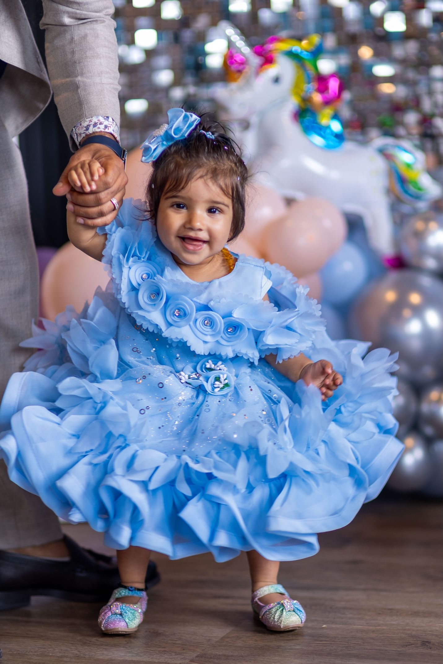 Smiling toddler girl in a blue ruffled dress with roses holds an adult's hand at a birthday party with balloons.