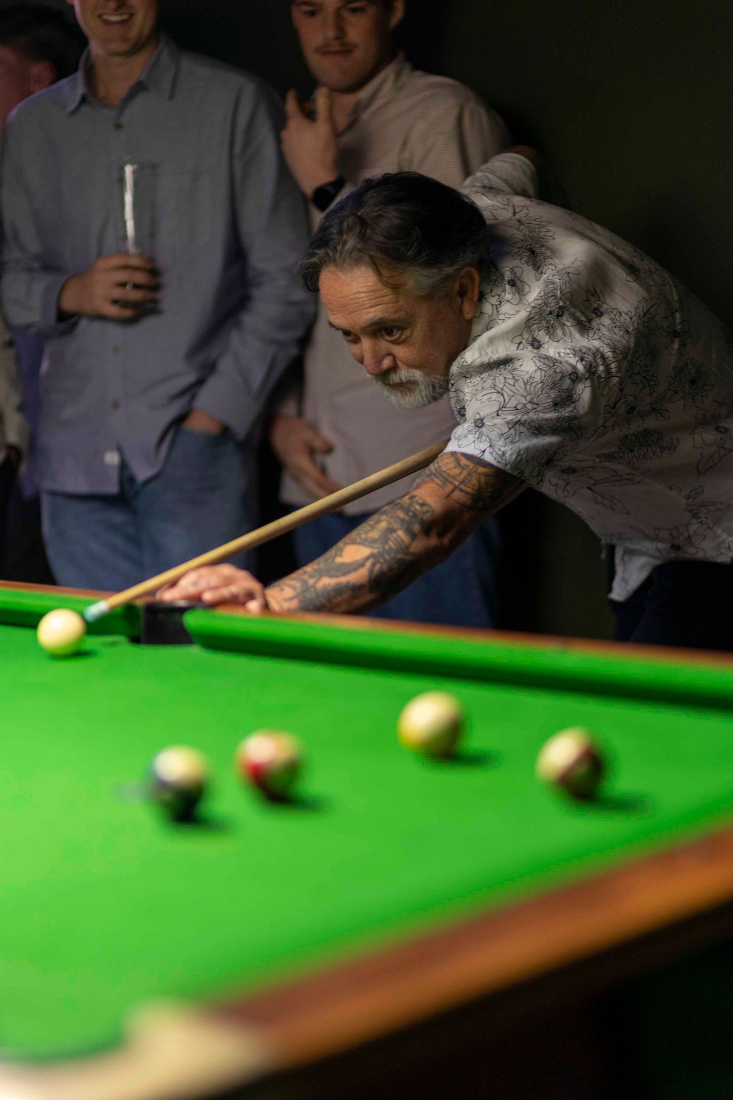 Tattooed man leaning over a green pool table taking a shot with a cue, watched by spectators in a dimly lit room.