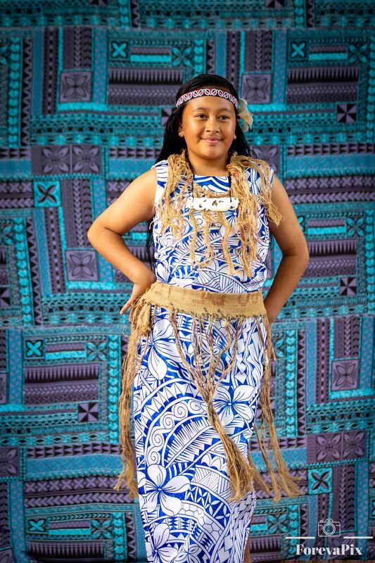 Young woman in traditional Polynesian attire with blue tribal print dress and woven accessories poses before tapa cloth.