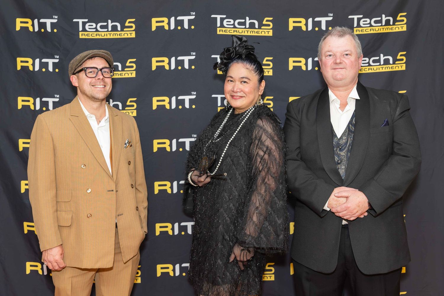 Three professionals pose together at an R.I.T. Tech 5 Awards event backdrop wearing formal attire.