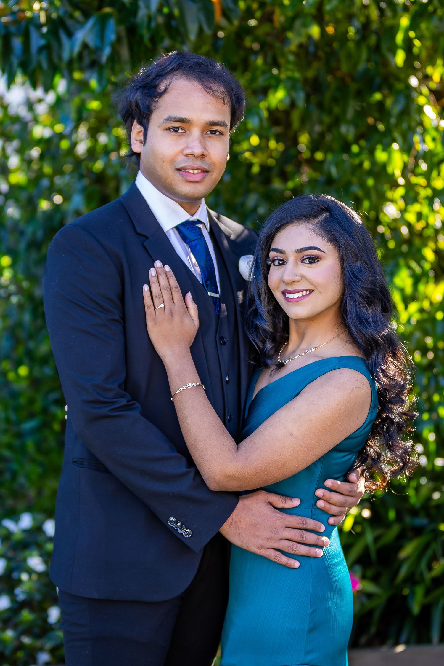 A young couple poses outdoors, man in navy suit, woman in teal dress, surrounded by lush green foliage.