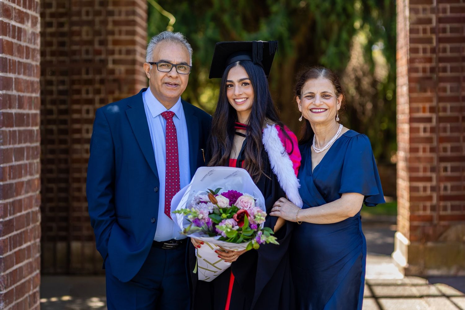 Graduate in cap and gown holds bouquet flanked by proud parents at brick university building.