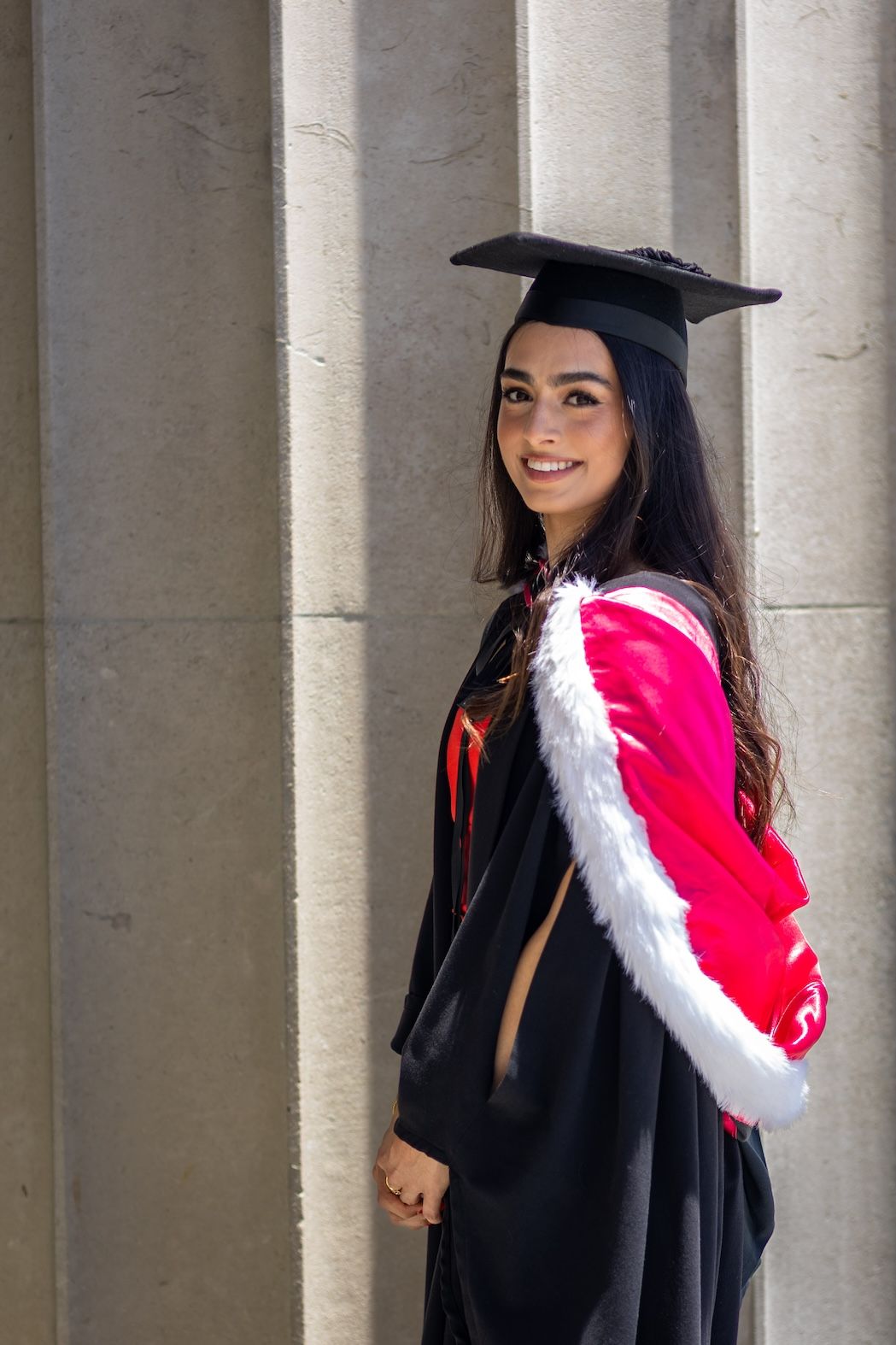 Smiling female graduate in black cap and gown with red and white hood poses between stone columns.