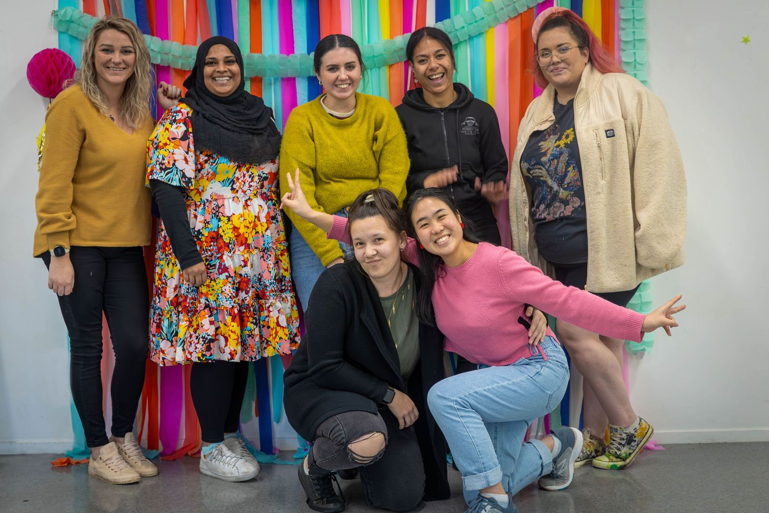 Group of seven diverse women smiling together in front of colorful fringe backdrop at indoor event.