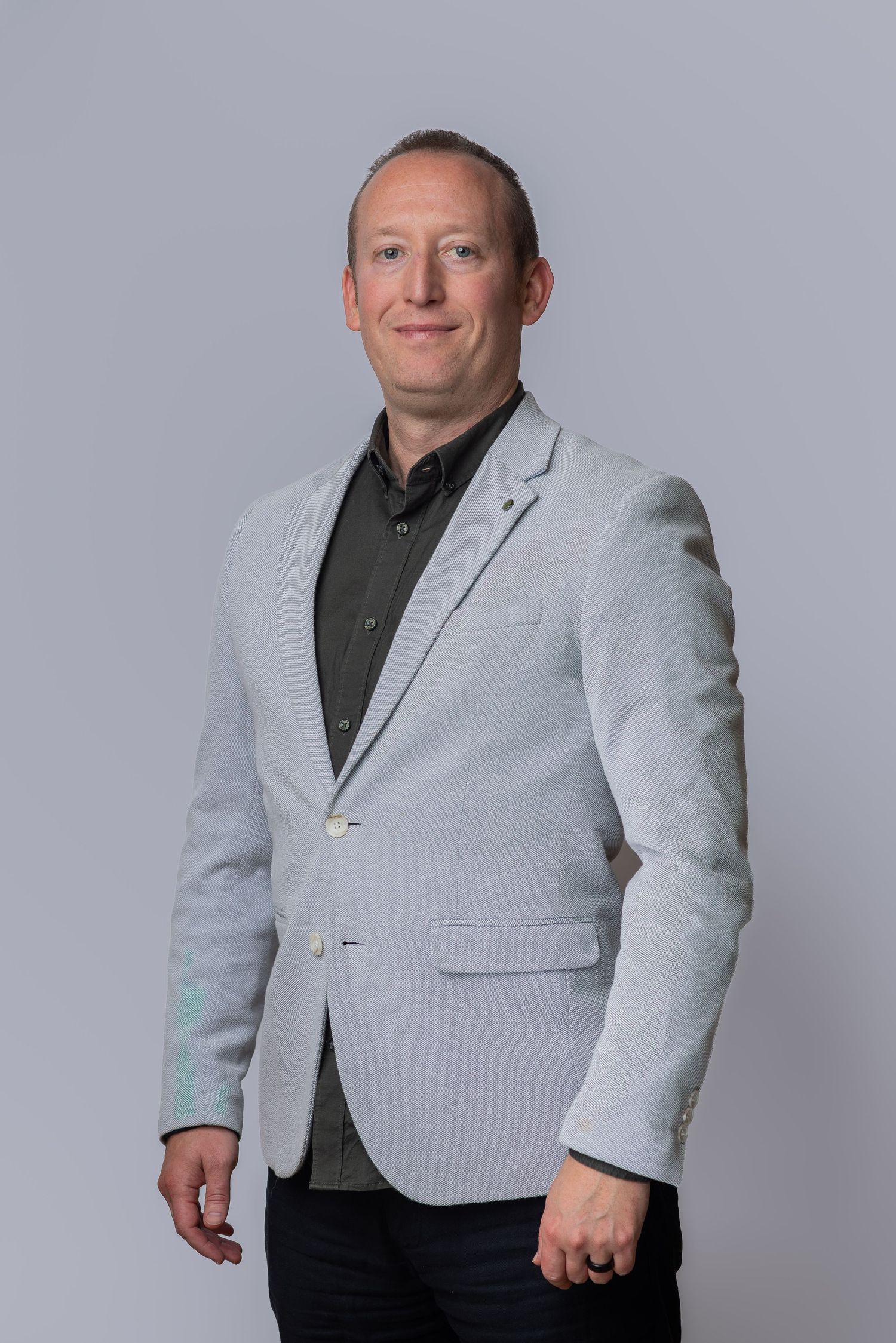 Professional man in light gray blazer and dark shirt posing confidently against a neutral gray background.