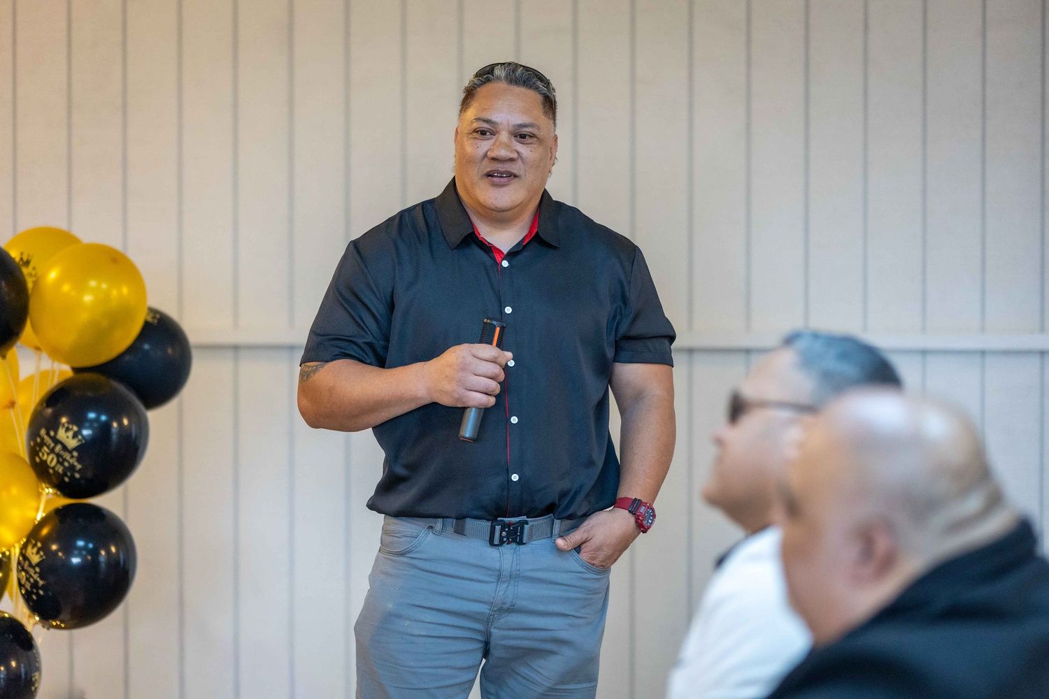 Man speaking at event with black and gold balloons, holding microphone in casual black shirt.