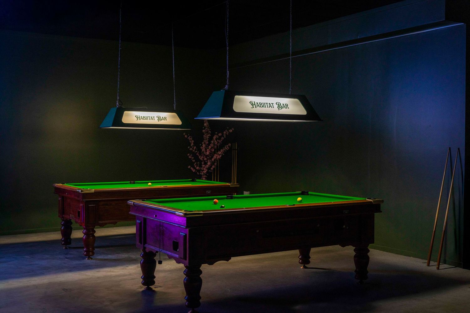 Two green felt pool tables with hanging Snooker bar lights in a dimly lit billiards room.