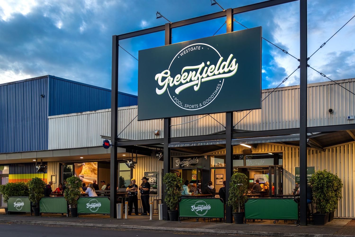 Exterior view of Broomfield restaurant with illuminated signage and green planters during dusk hours.