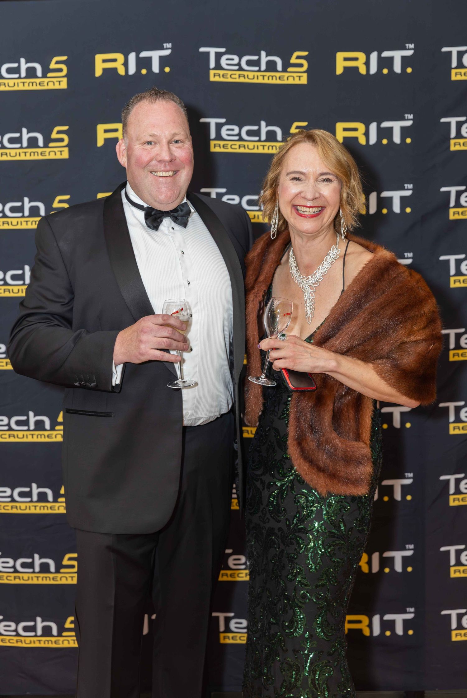 Elegantly dressed couple posing at Tech5 Recruitment and R.I.T. event, man in tuxedo and woman in green gown with fur wrap.