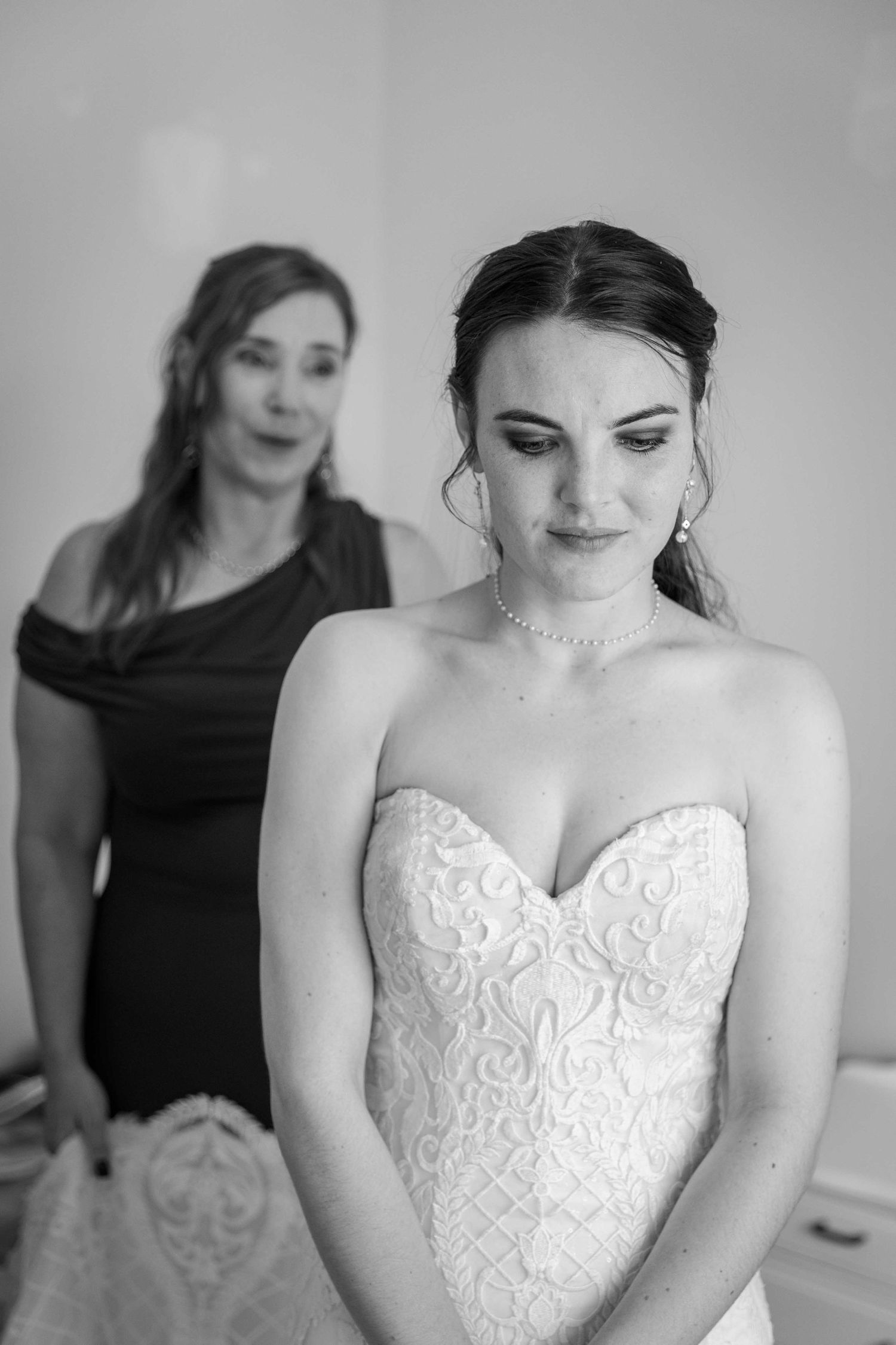 Black and white photo of bride in strapless lace gown with woman helping her prepare in background.