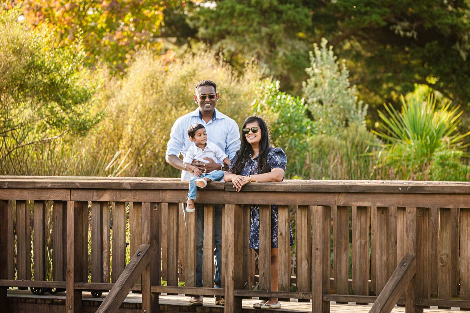 Family of three posing on a wooden bridge surrounded by lush green trees in an outdoor park setting.