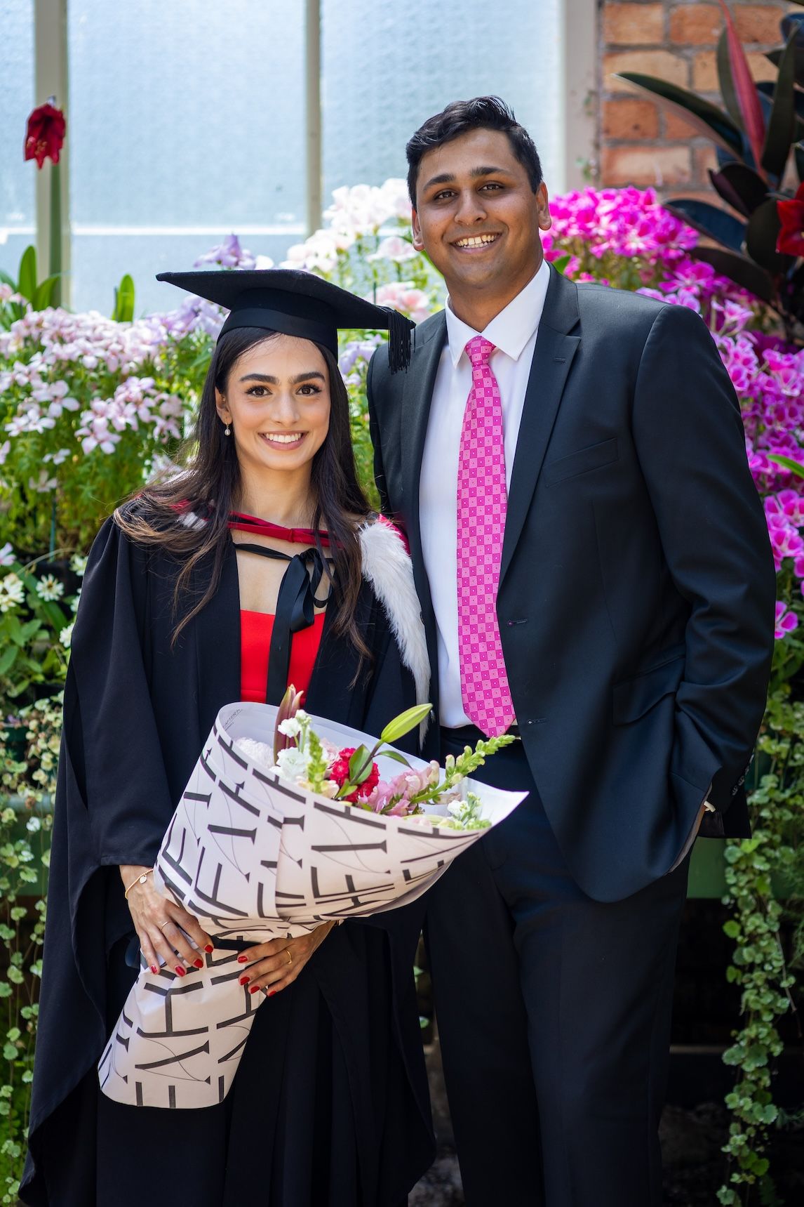 Smiling graduate in cap and gown holds bouquet beside man in black suit at graduation.