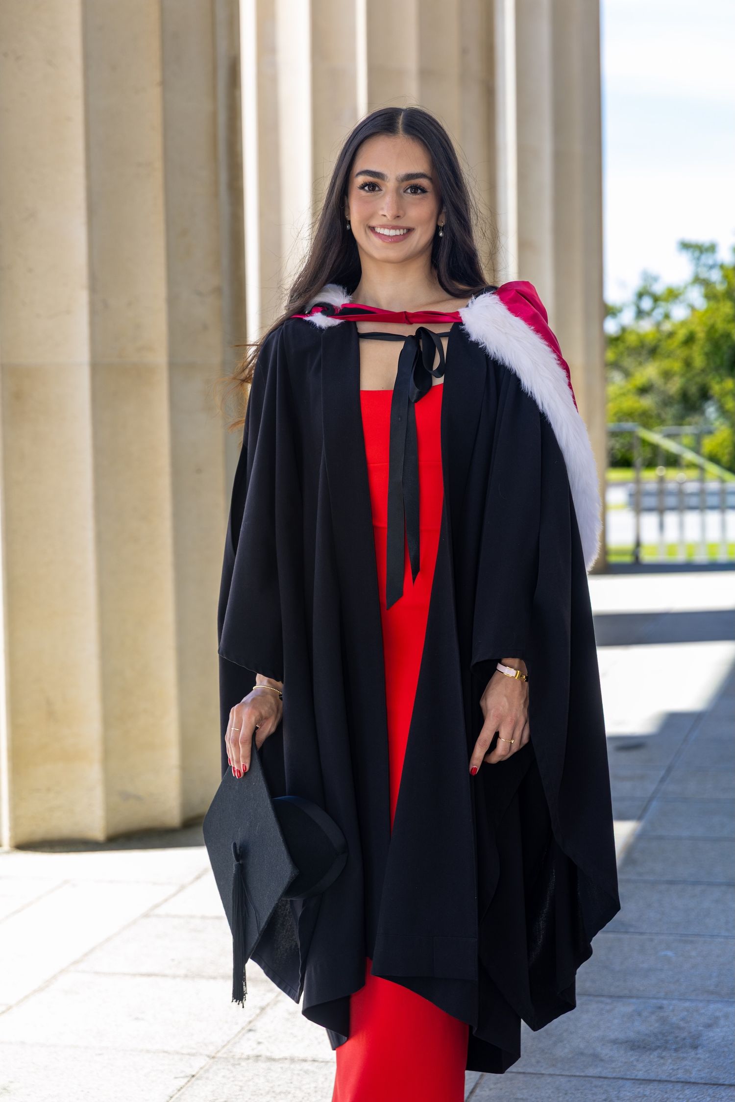 Smiling graduate in black gown with red hood holding mortarboard cap, wearing red dress, posing by stone columns.