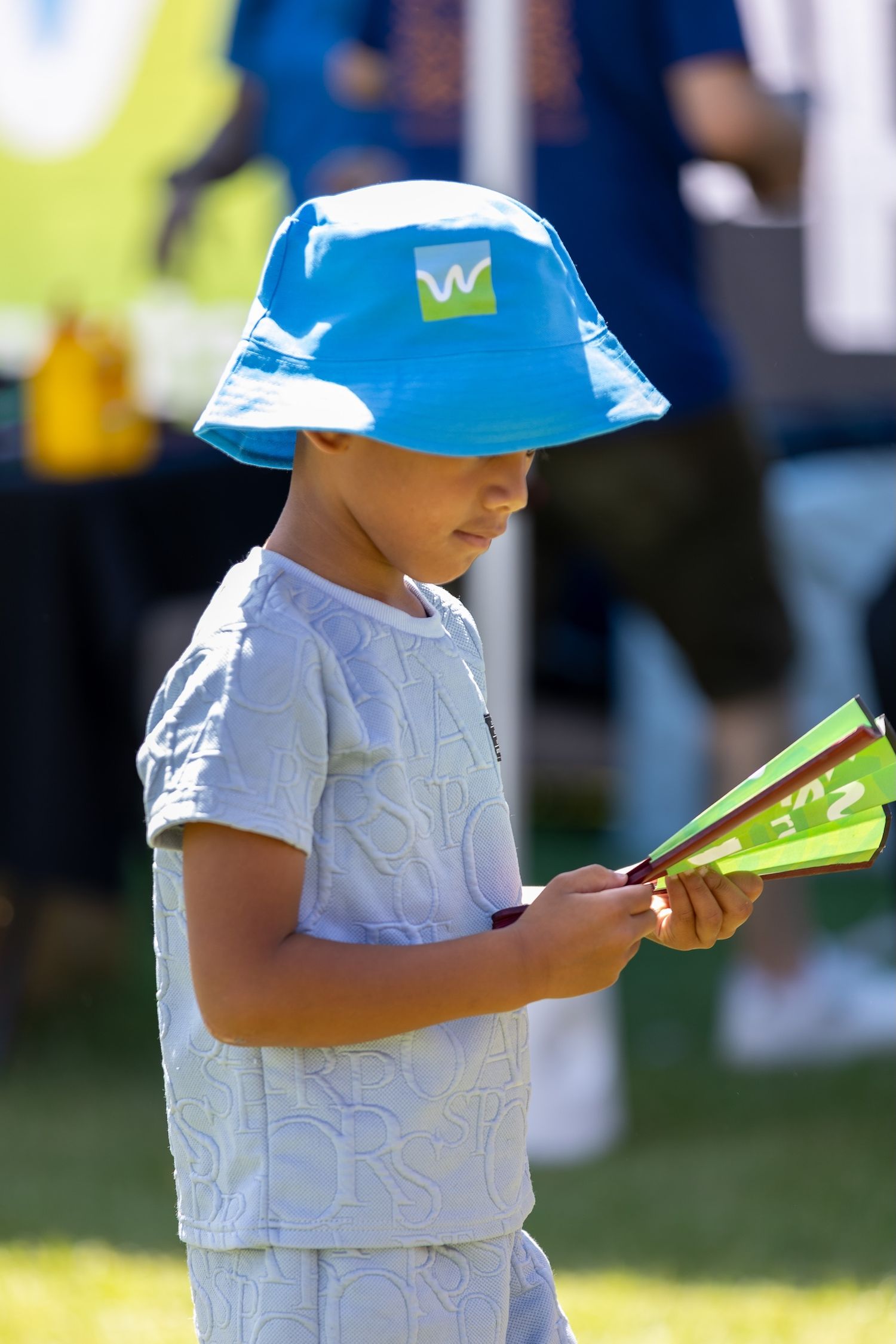 Child in blue hat and white shirt holds a green folding fan at outdoor event.