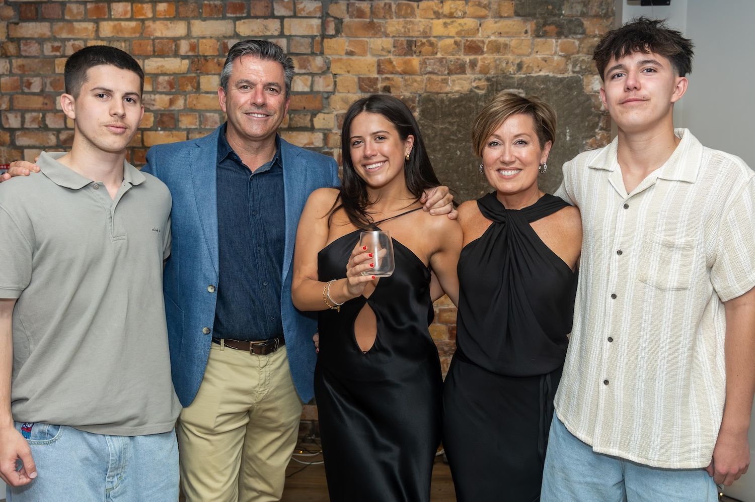 Family of five posing together smiling at a celebration event against a brick wall backdrop.