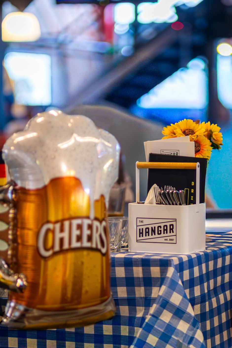 A beer glass and napkin holder with sunflowers on a blue checkered tablecloth at a restaurant table.
