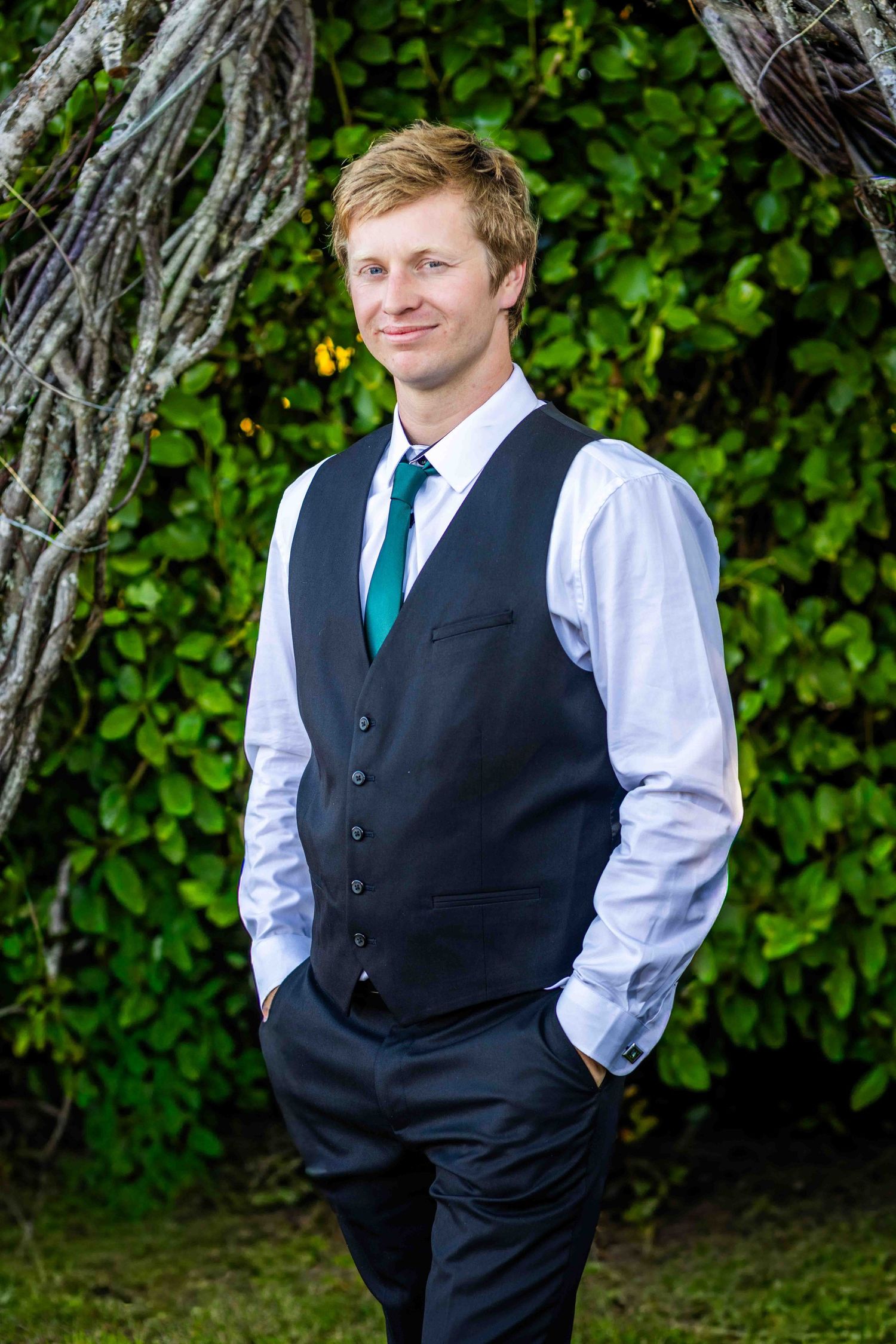 Smiling groom in dark vest, white shirt, and teal tie standing outdoors in front of lush green foliage.