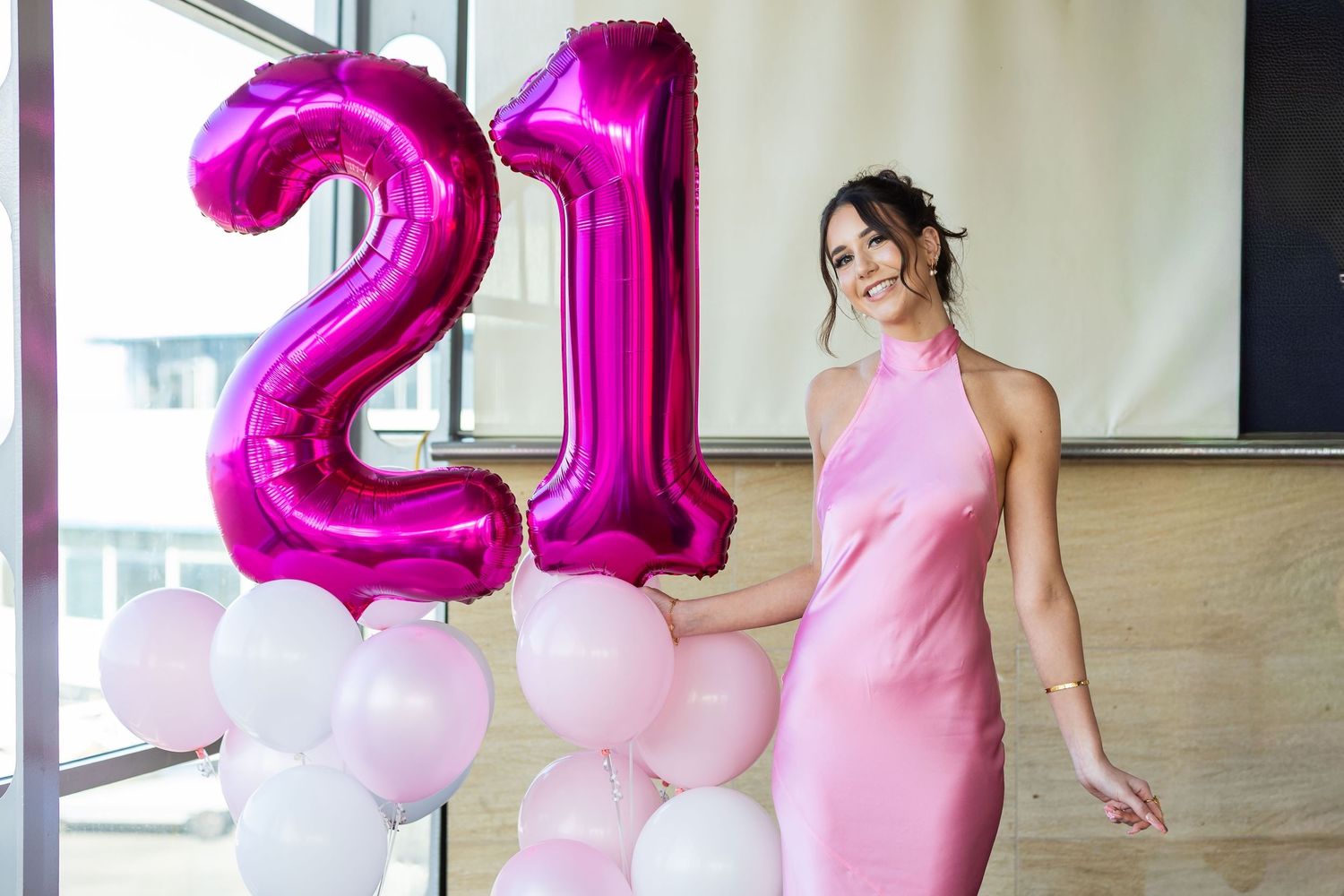 Young woman in pink dress smiling beside large magenta number 21 balloons at a birthday celebration.