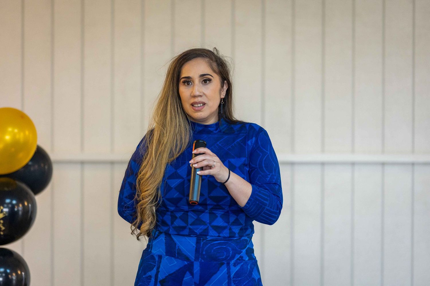 Woman in blue patterned dress speaking at event holding microphone with black and gold balloons in background.