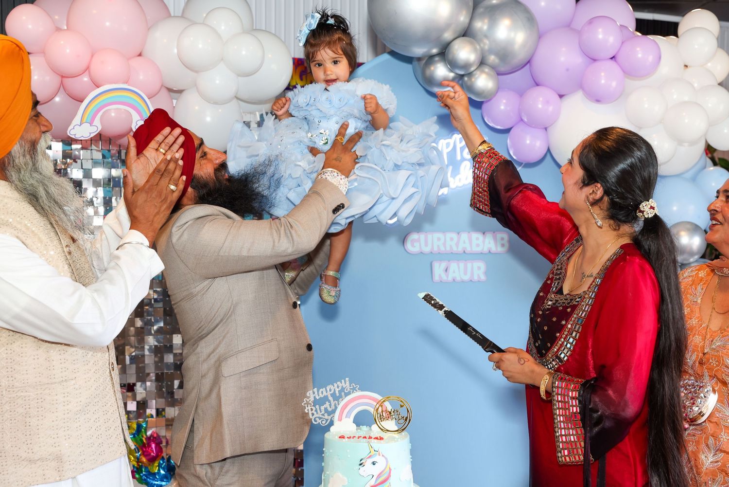 Family celebrating a baby girl's first birthday party with balloons and decorations at a colorful event.