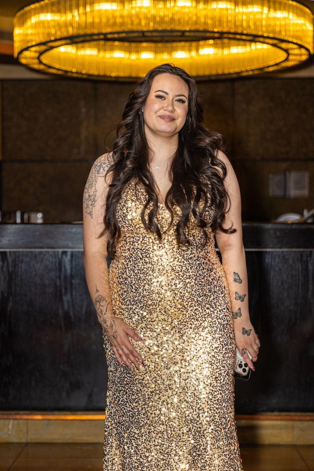 Woman in gold sequin gown with tattoos poses under chandelier in elegant venue.