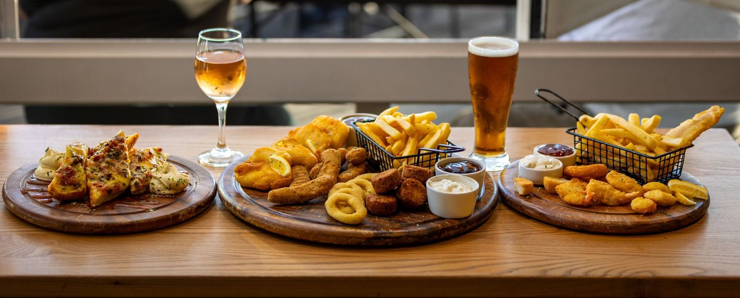 Assorted pub food and drinks including burgers, fries, cocktails and beer arranged on a wooden bar counter.