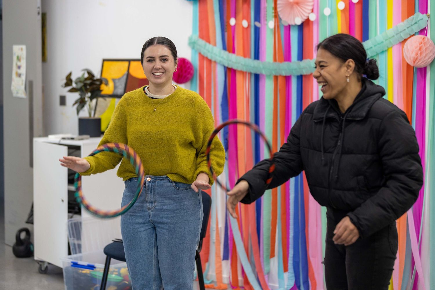 Two women smiling and playing with hula hoops in a colorfully decorated room with rainbow streamers.