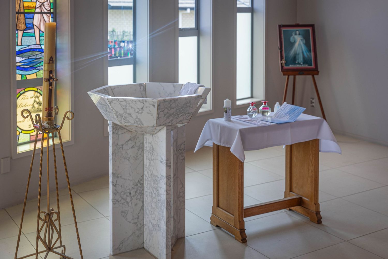 Marble baptismal font beside a white-draped altar table in a bright chapel with stained glass windows.