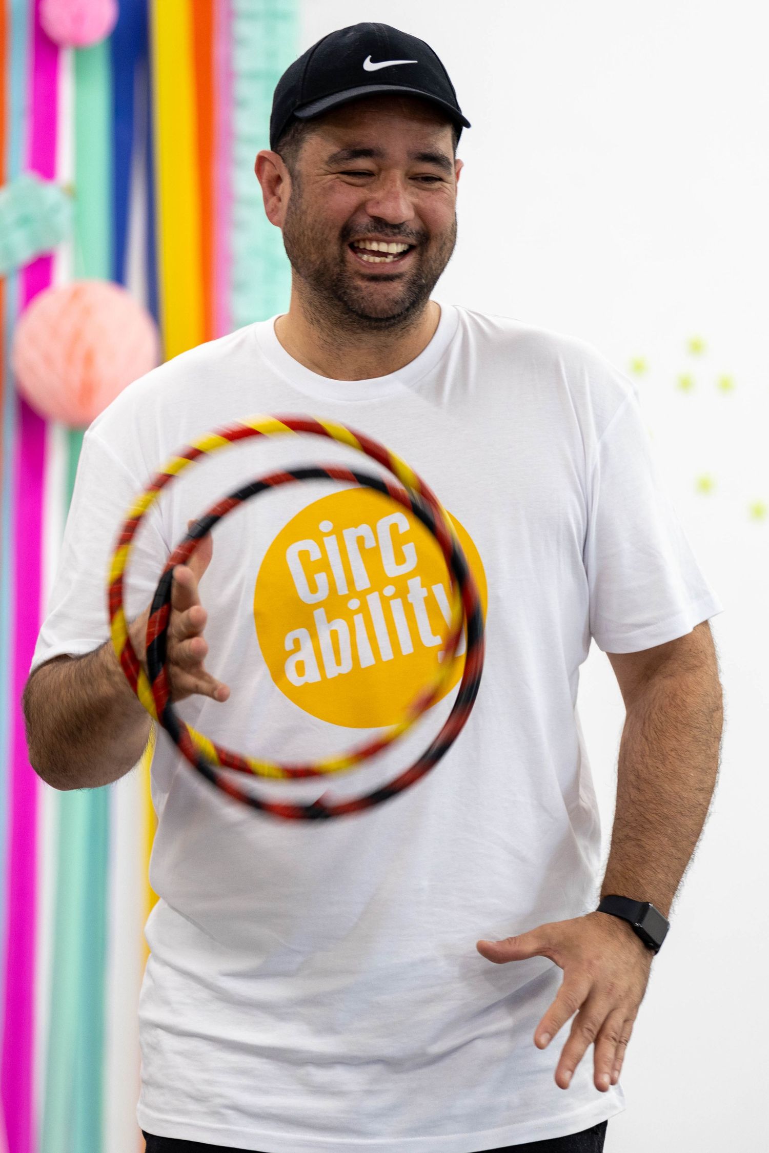 Smiling man in white CircAbility t-shirt spinning a colorful hula hoop, wearing a black Nike cap.
