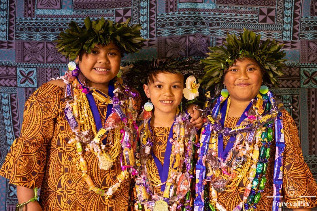 Three children in traditional Polynesian attire with floral leis and leaf crowns smile at a cultural celebration.