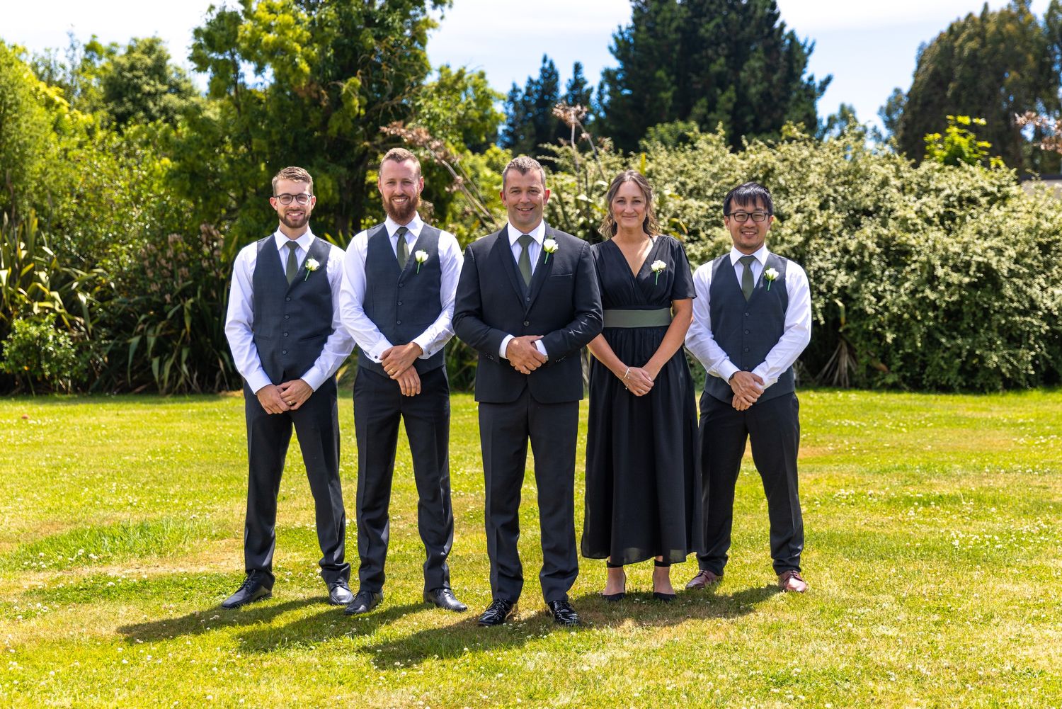 Wedding party of five posing outdoors on green lawn with lush garden backdrop on a sunny day.