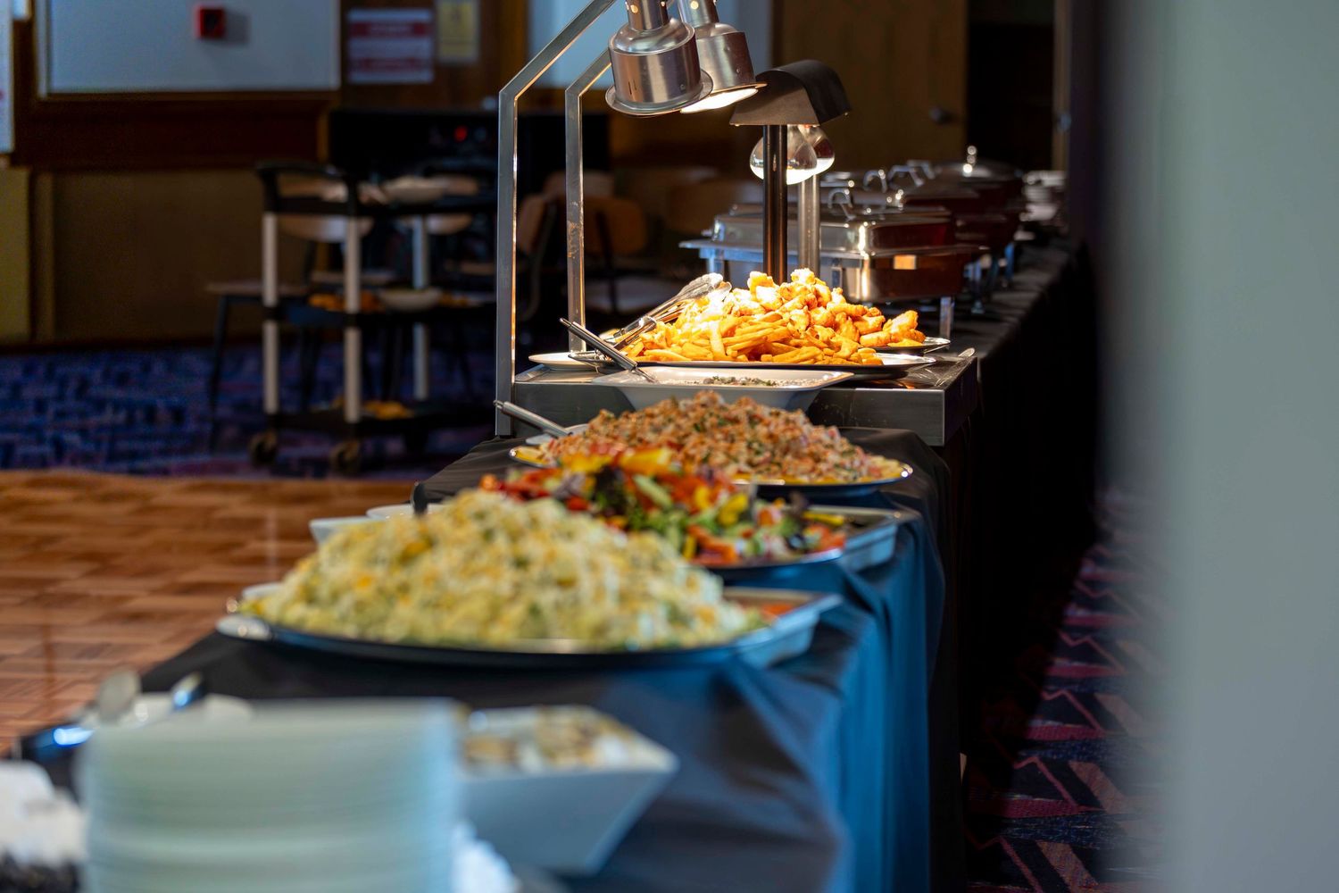 A buffet line with heated serving stations displaying various dishes on blue tablecloths under warming lamps.