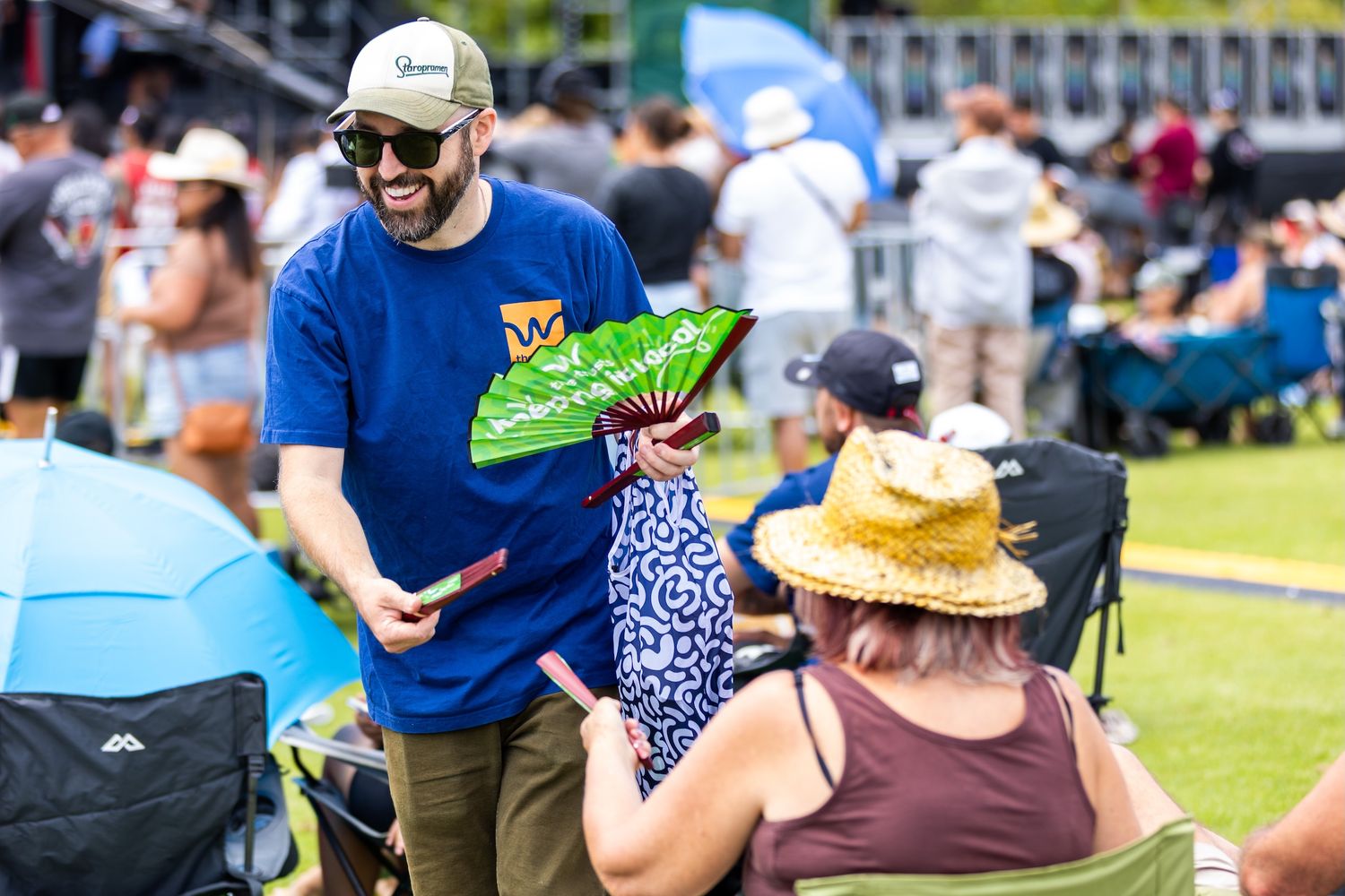 Festival-goers relaxing on grass with umbrellas and tents visible in background.