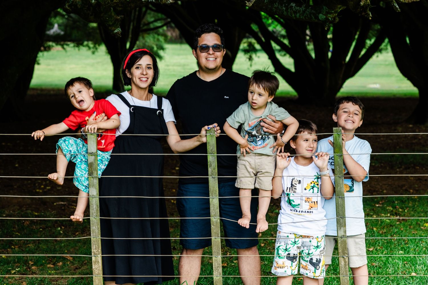 Family of six posing outdoors near a wooden fence with lush green trees in the background on a sunny day.