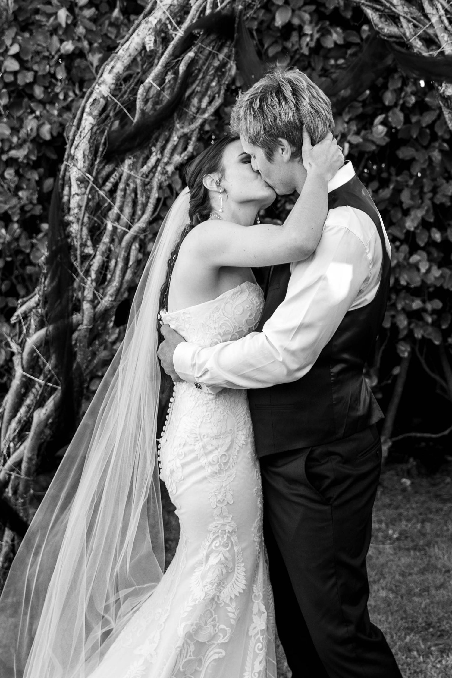 Black and white photo of a bride and groom sharing a romantic kiss outdoors near a vine-covered tree.