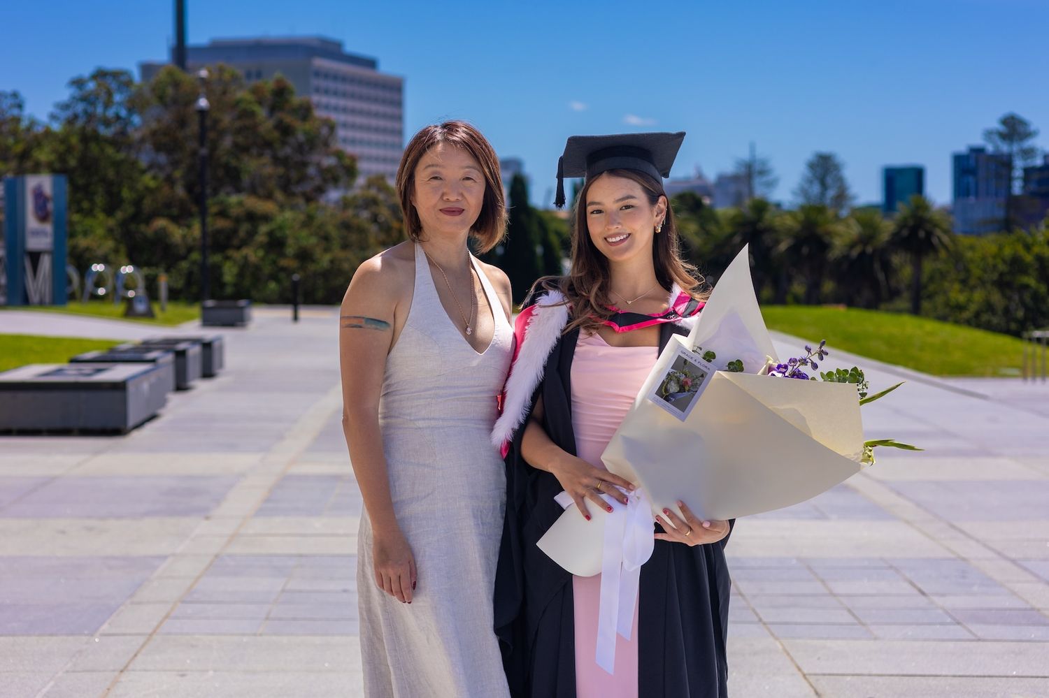 Two women pose outdoors at a graduation ceremony, one wearing cap and gown holding a bouquet on a sunny day.