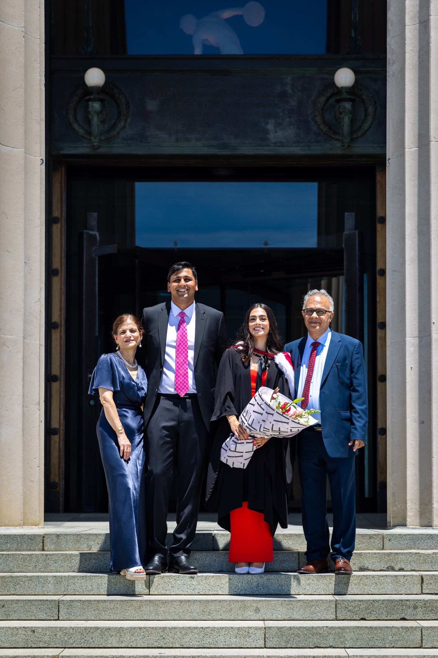 Four people pose on steps outside a building, one wearing graduation gown holding flowers, celebrating a graduation day.
