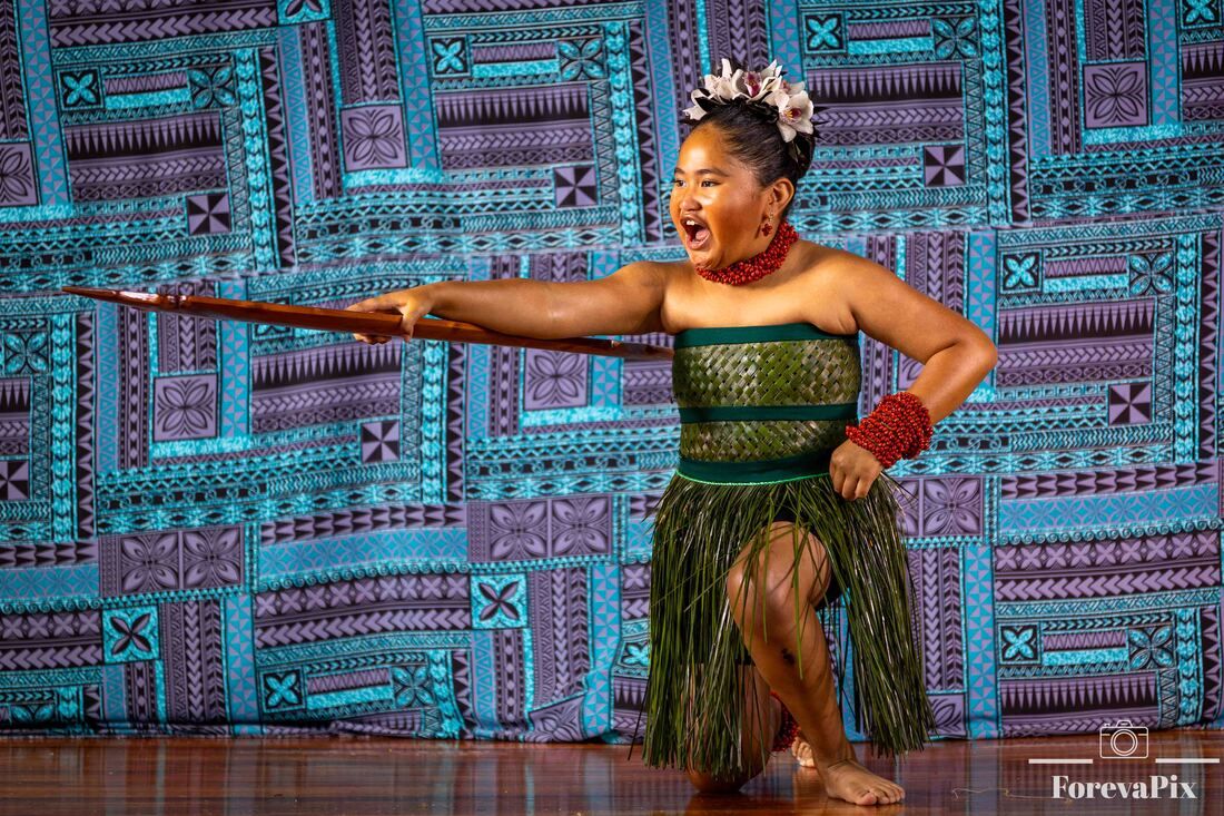 Polynesian dancer in traditional green costume performs with a spear against a colorful tapa cloth backdrop.