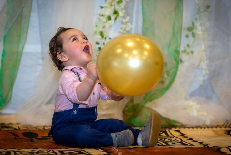 Joyful baby girl holding a gold balloon, sitting on a rug with white curtains in the background.