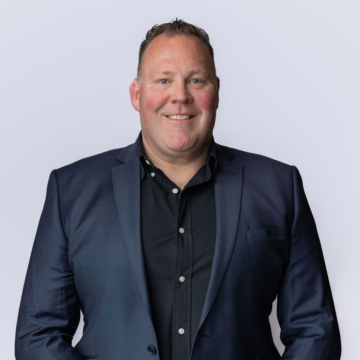 Professional headshot of a smiling man in a navy blue blazer and black shirt against a light gray background.