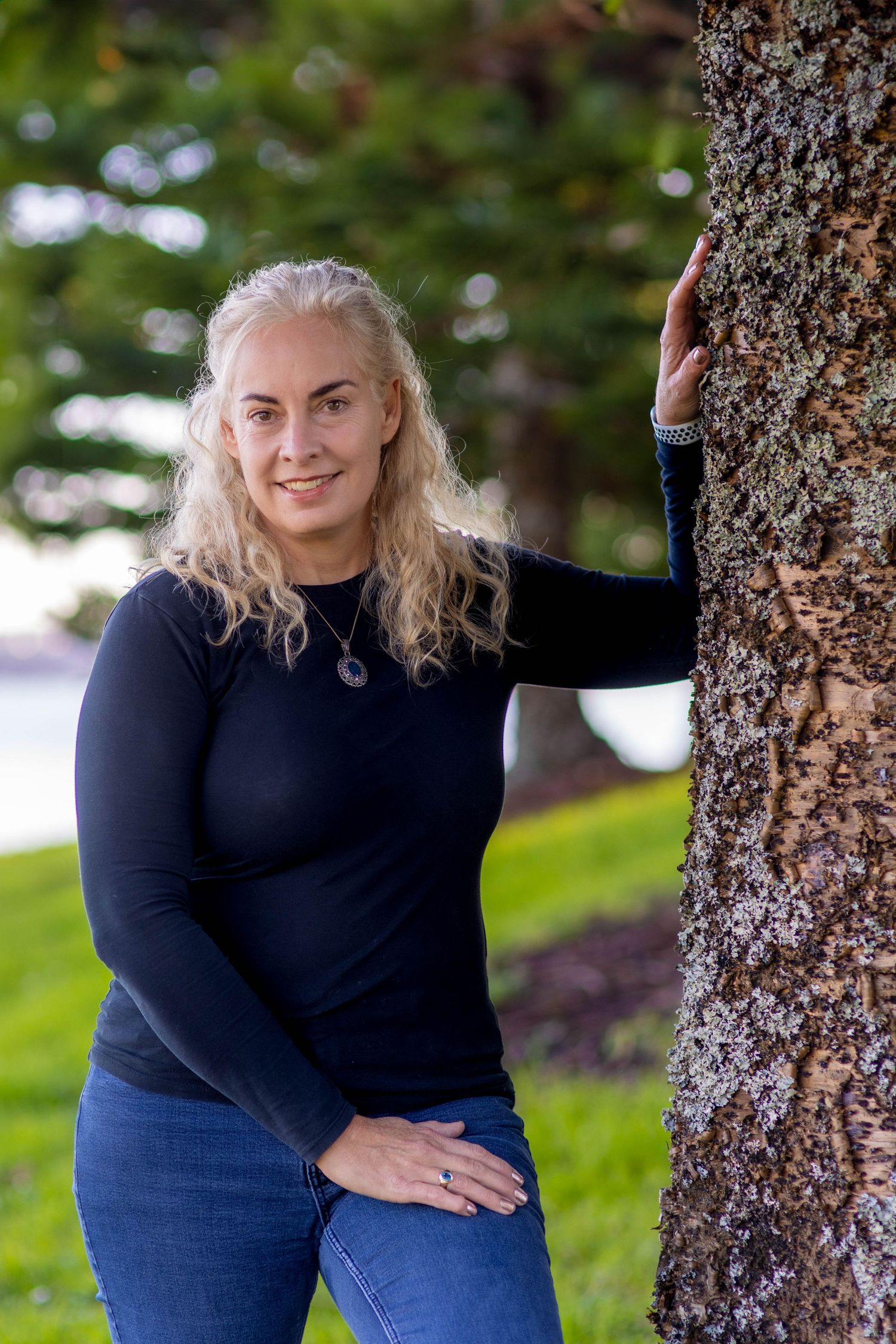 Woman with blonde curly hair in black top and jeans leaning against a tree in a park.