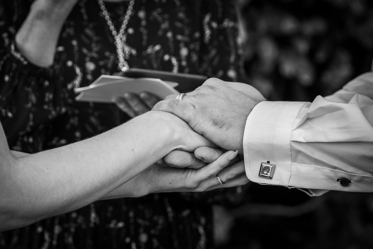 Black and white photo of two people holding hands, with cufflinks visible, during a wedding ceremony.