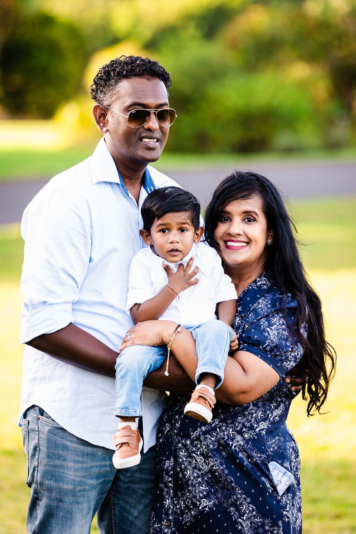 Happy South Asian family with parents holding young toddler son, smiling outdoors in a green park setting.