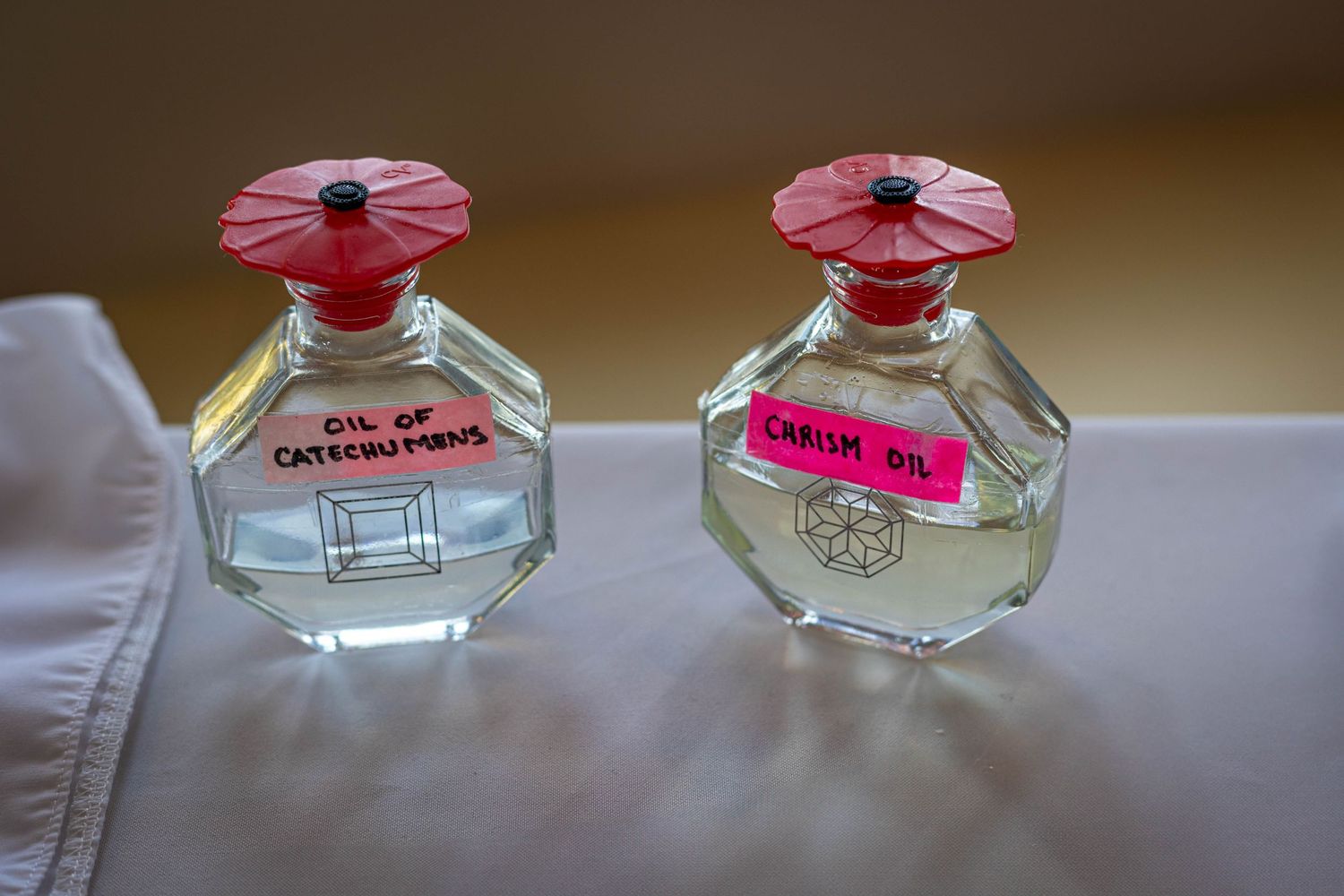Two glass perfume bottles with red caps labeled Oil of Catechumens and Chrism Oil on a white cloth surface.